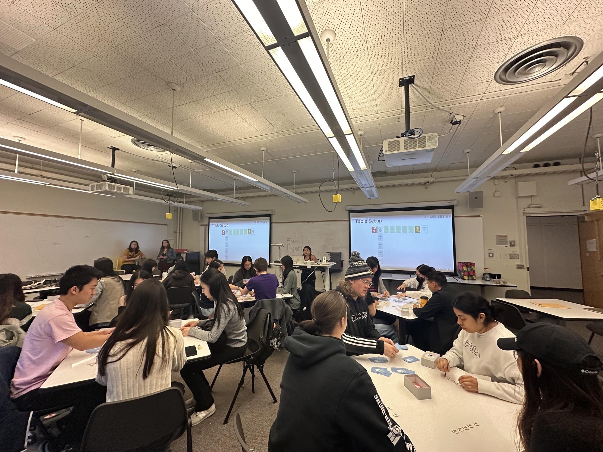 A full classroom of roughly thirty students playing the finished game in teams, with table setup instructions projected on two screens